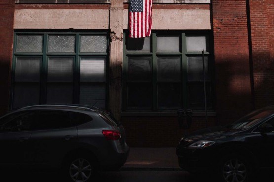 street photo of brick city building with two cars parked in shadows and american flag hanging from building in the sun above