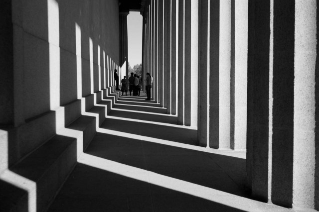 black and white outdoor shot of building pathway with light streaming through large pillars on the right