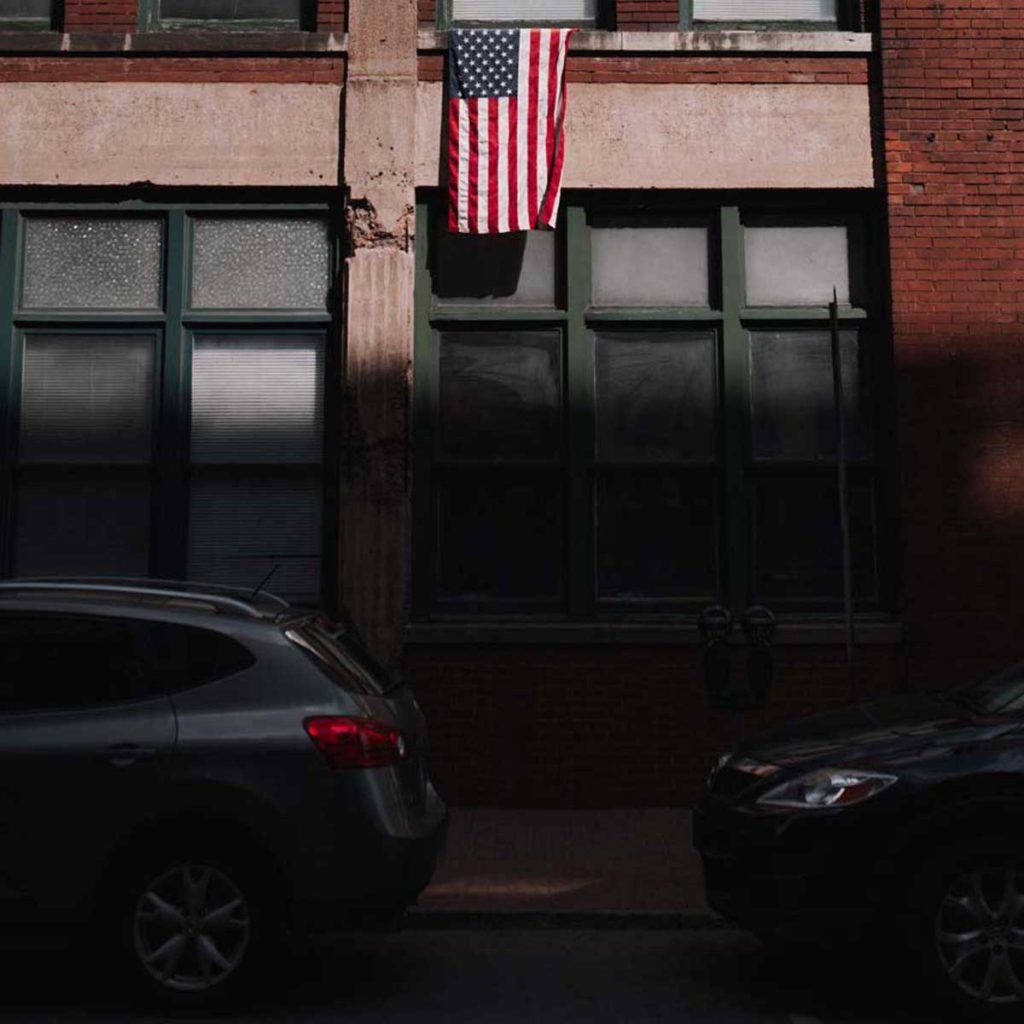 street photo of brick city building with two cars parked in shadows and american flag hanging from building in the sun above